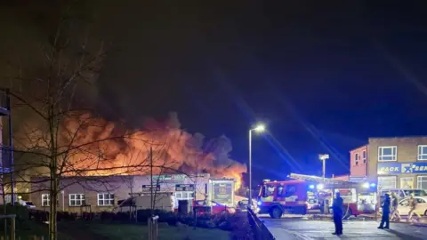 Paul King A picture of a well-developed fire in a single-storey industrial building at night. An orange glow comes from the building and it is dark as a fire engine is parked in front of it.