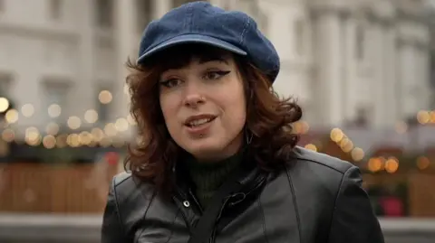 Serena Kaos is pictured in Trafalgar Square, wearing a blue cap and black jacket, with the Christmas market blurred in the background.