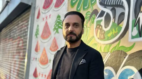 Abdul Malik, wearing a navy jumper and jacket. He is looking at hte camera and there is colourful street are on the wall behind him. 