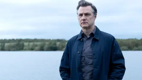 BBC/House Productions/Sam Taylor A man in a dark blue jacket with a blue shirt underneath stands in front of a body of water, there are trees and fields in the further distance