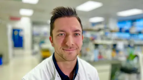 BBC/Martin Giles Damien Caubriere smiles at the camera. He has short, spiky brown hair and is wearing a white lab coat over a dark-coloured shirt. He is standing in a laboratory.