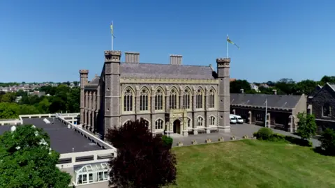Victoria College in Jersey. The main building is made from grey stone and has large church-like windows. The building has turrets on the corners. A large green lawn is in front of the building. It is a sunny day.