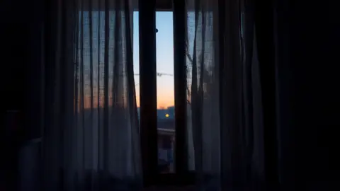 Getty Images Thin curtains at a window in a very dark room. Outside through a gap in the curtains the sun is setting, giving the sky an orange glow.
