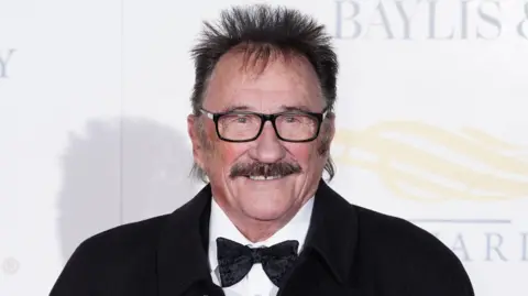 Paul Chuckle at a red carpet event. It is a close crop of his face. He has a brown moustache, brown hair sticking up around his head and black square glasses. He is smiling and wears a white shirt, black bowtie and black jacket.