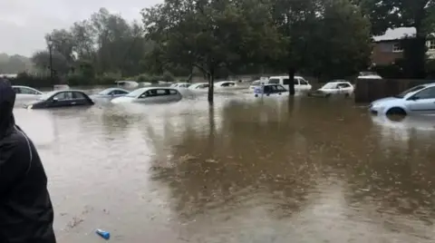 Hazik/Weather Watchers A flooded area with a more than ten cars submerged under water, with water up to the door handles of the cars. The water is brown and raindrops are visible on the water's surface. 