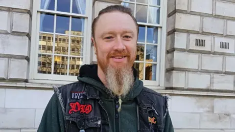 Kenneth McClernon has short brown hair slicked back and a long ginger/grey beard. Her is wearing an armless denim jacket with patches on heavy metal groups on it and a green hoodie 