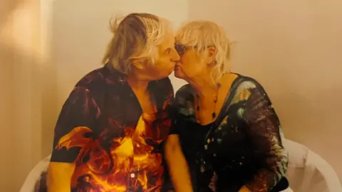 A man and a woman, both in dark-coloured tops with a flowery design are sat down in a beige room and leaning in to each other in a kiss