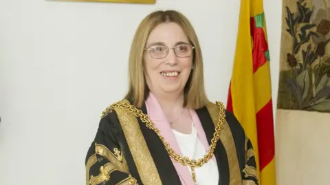 Stuart Walker Mayor Jeanette Whalen wearing the existing robe at her mayor-making earlier this year. She has long blonde hair and is wearing glasses. She has a gold chain around her neck and is wearing a black robe decorated with gold embroidery