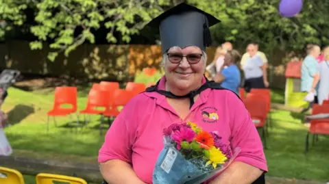 Sheila Dwyer smiles as she holds a bouquet of flowers with a graduation mortar board on her head in a garden