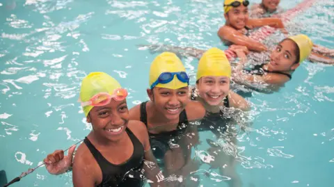 Getty Images A group of school girls in yellow swimming hats, some with goggles on their heads, in a swimming pool.