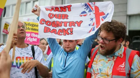 An image of an outdoor protest. A man in the middle, wearing a blue jumper, holds a sign above his head reading "want our country back", with an image of Britain and Northern Ireland coloured in the colours of the union flag. A man to his left is shouting with his mouth wide open, while a man on his right is wearing a t-shirt with "love" in rainbow colours on the front. 