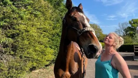 Roo Fox, who has blonde hair and is wearing a light blue top, is on the right of the picture and her horse Frog is on the left. They are standing on a path with a hedge and trees in the background.