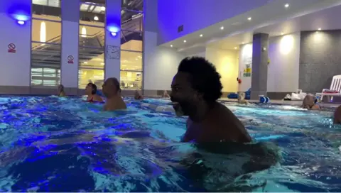 Alvan in a swimming pool with other people closeby in an aqua aerobics class.