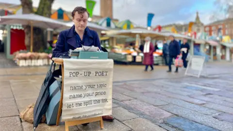 Martin Giles/BBC Rodney-Jones is sitting at a small table on the paving slabs at Norwich Market, with the covered market stalls behind him out of focus. Historical buildings can be seen in the background, including City Hall. He has a turquoise typewriter and a cloth draped over his table which reads: POP UP POET. NAME YOUR TOPIC. NAME YOUR PRICE. LET ME SPILL THE INK. 