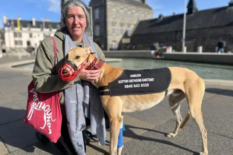 A woman with grey hair kneels beside a greyhound, which has a black rug on with Scottish greyhound sanctuary branding. There is a pool of water and stone buildings in the background. 