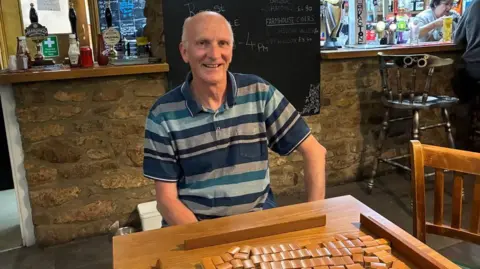 Mark Trenchard set up Shipham's Mahjong club. He is a slim, middle-aged white man who is wearing a blue striped top. He is sitting in a pub with a Mahjong table in front of him. 