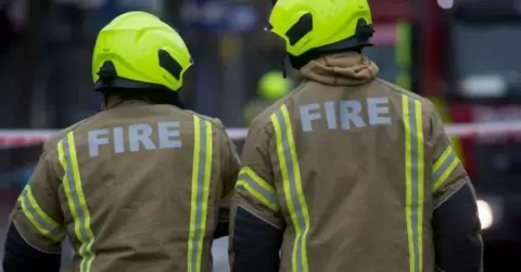 Getty Images Two firefighters stand with their backs to the camera, wearing yellow fire jackets and helmets. Their heads are tilted towards each other. 