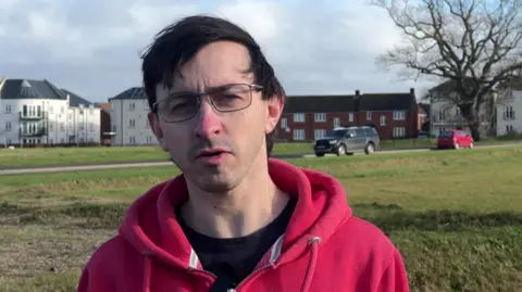 Jordan Crawford ina red hoodie standing on a large area of open grass, with a road and houses and blocks of flats in the distance behind him.
