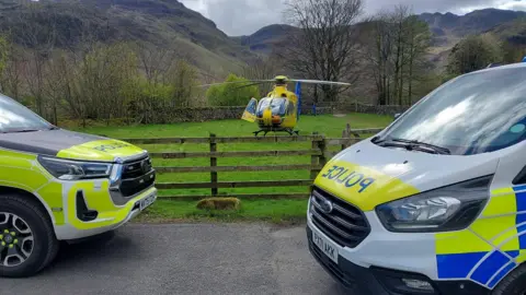 Two yellow, blue and white police cars parked facing each other in front of a field. In the field there is a yellow and blue helicopter with one of its doors open. There are mountains behind the field and the sky looks grey.