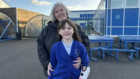 Zoey is in her blue school uniform and is smiling at the camera. She has brown hair and a fringe. Part of he bionic arm can be seen as the sleeve of her cardigan has been pushed up. The bionic arm has a white casing and black outlining. Her grandmother is hugging Zoey from and has her arms wrapped around her waist. The grandmother is wearing a black puffer coat and is smiling into the camera. Behind them is Zoey's school, which is a blue block with rectangular windows. To the left of them are bike sheds with bike racks in them and there are blue outdoor metal tables and benches on the right. 