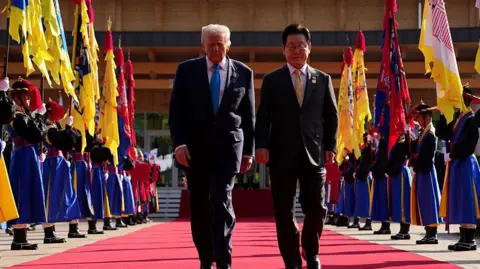 Getty Images President Donald Trump walks with South Korean President Lee Jae Myung down a red carpet flanked with mascots carrying flags 