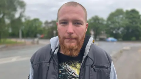 John stands on the side of the road, wearing a t shirt and hooded jacket, looking straight into the camera.