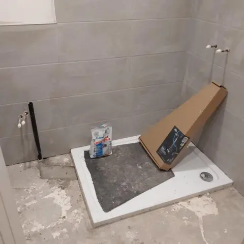 Peter McCombe Unfinished bathroom with concrete floor, shower tray, but no shower cubicle, and tap points coming out of the wall, but no taps. The walls are tiled a grey colour that looks unfinished. The shower tray has a cardboard box on it, and a packet that looks like grout or other building materials, along with a section of black matting or underlay.