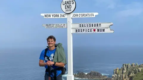 Man stood by a sign at Land's End which shows distances to different locations, with the miles it would take to get there. The sign has New York and John O'Groats on it and also a sign reading "Salisbury Hospice/Mum"