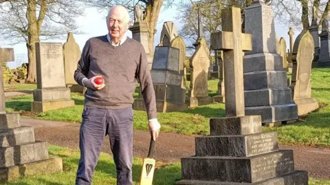 Ian Lockwood A man stood in a graveyard, holding a cricket bat and red cricket ball. There are a number of headstones and statues around him with stone crosses. There is also a path behind him.