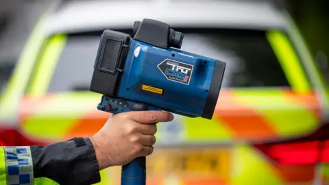 PSNI A police officer holds a speed detection camera. a police vehicle is in the background