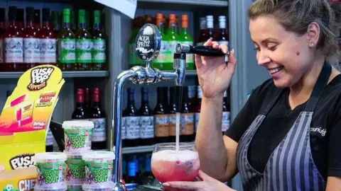 Ashleigh Morley Ashleigh smiles as she pours red juice from a tap. She has fair hair worn in a bun on top of her head. She wears a black t-shirt under a blue-striped apron. The shelves behind her are stacked with cordials.