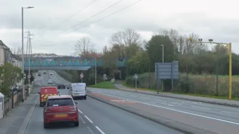 General view of a road. Cars are travelling along the road. Trees and houses line either side of it. Power lines are visible above the road.