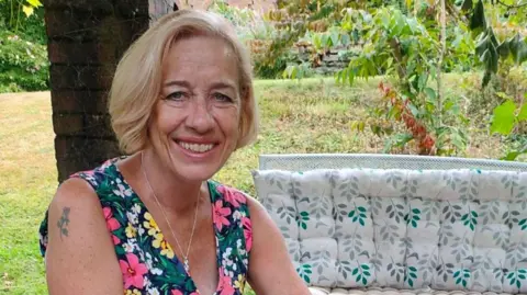 Angela Tilley Woman with short blonde hair and a floral dress smiling at the camera sat on a bench 
