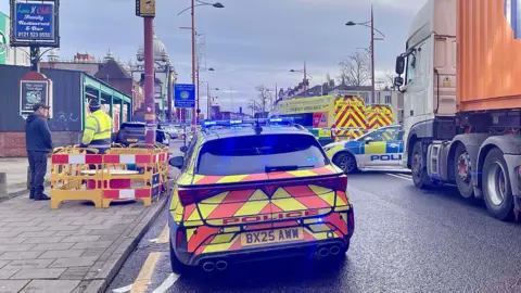 Soho BID A police car with it's blue lights on parked on a cordoned off road. There are two amublances and an another police car parked beside blue and white police tape.