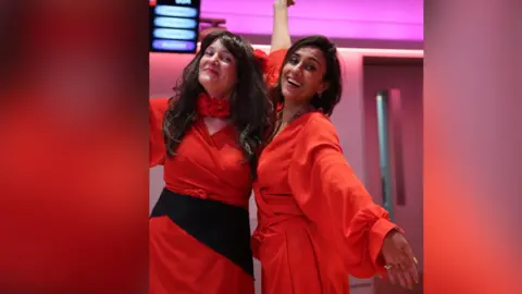 Two women embrace and pose for the camera. They are wearing red dresses like Kate Bush.