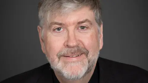 A portrait style picture or Tim Barker wearing dark clothing. He has short grey hair and a short grey beard. 