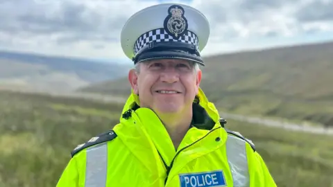 Insp John Mitchell wears a hi-vis jacket, which has a blue badge reading police in blue and white. He wears a white hat with the force's emblem. He stands on a green hill next to a road.