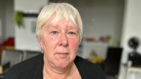 Sue Cheetham is staring directly at the camera. She has short hair and is wearing a black cardigan.
