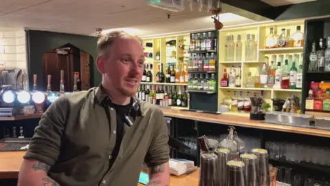 Josh Bate in a black T-shirt and olive shirt, leaning on the bar of Oriels