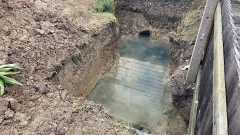 A hole is dug into the ground and filled with water. It is next to a fence.