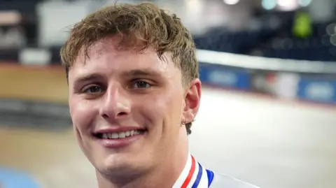 Matthew smiles at the camera. He has short brown hair and is wearing the Team GB race suit with medals around his neck.