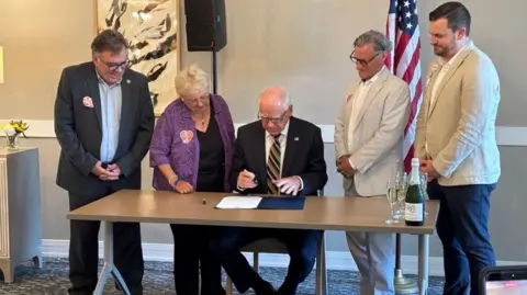 Amira Choice and Ebenezer Senior Living Minnesota Gov. Tim Walz wears a dark suit and striped tie as he sits at a table, an open folder in front of him, holding a pen as he prepares to sign legislation; Anita LeBrun wears a purple blazer, black shirt and black pants as she stands next to him. Three other men watch the signing with am American flag in the background