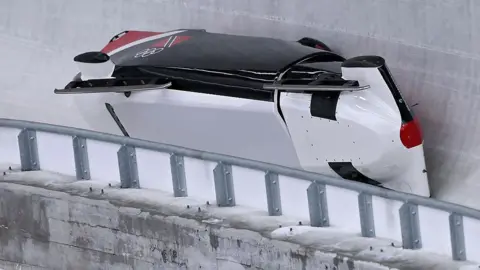 Getty Images A bobsled tipped onto its side while sliding along the curved wall of an icy track. The sled is mostly white with black and red sections. Metal safety barriers run along the lower edge of the track in the foreground.