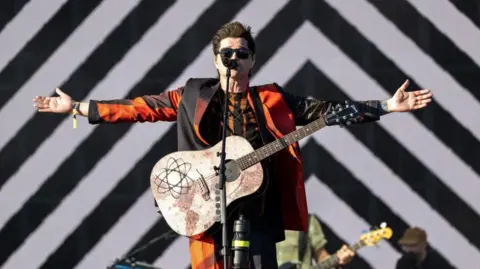 A man in a red suit with an acoustic guitar with his arms spread wide at a mic