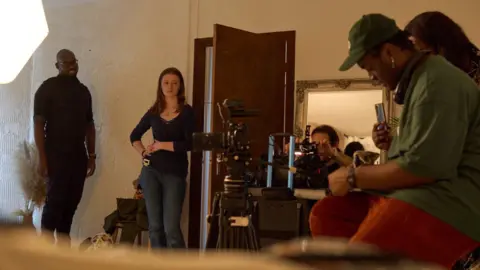 Jimmy Osayamwen The crew on set of Rice at Home. There are multiple people in a room with a brown wooden door. A black man to the right of the photo, in a green shirt is controlling a camera. Various people are standing around, concentrating on a focal point off screen.