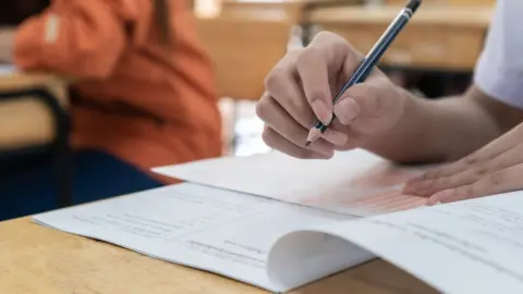 A close-up of a girl's hand is seen writing with a pencil on a test paper in what looks like an exam hall in a school