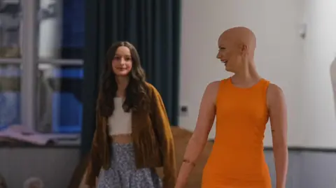 Tenovus Cancer Care Enlli Thompson taking part in a fashion show for the charity, Tenovus Cancer Care. Her head is shaven and she is wearing a short sleeve orange dress. To her right is another young girl who is also taking part in the fashion show. She is wearing a brown jacket, white crop top and a purple floral skirt.