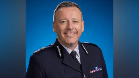 A close-up photo of the acting chief constable of West Midlands Police. He wears a black police uniform and stands in front of a blue background, looking into the camera.
