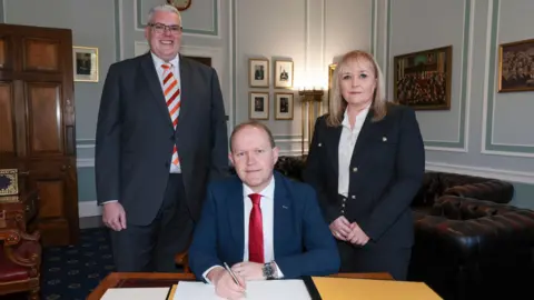 A man sitting at a desk he is holding a pen wearing a red tie and navy blazer. Behind him is a man and woman, the man is wearing a black suit and red and white tie and the woman is wearing a black suit and white shirt.