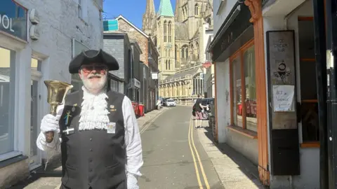 BBC A town crier in ceremonial dress including a black hat and white laced shirt holds a gold bell and smiles at the camera in the street. 